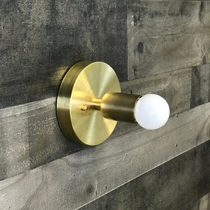 May include: A gold-toned wall sconce with a round base and a single light bulb. The sconce is mounted on a gray wood wall.
