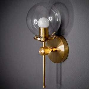 May include: A gold wall sconce with a clear glass globe shade and a single light bulb. The sconce has a long, thin arm that extends out from the wall.