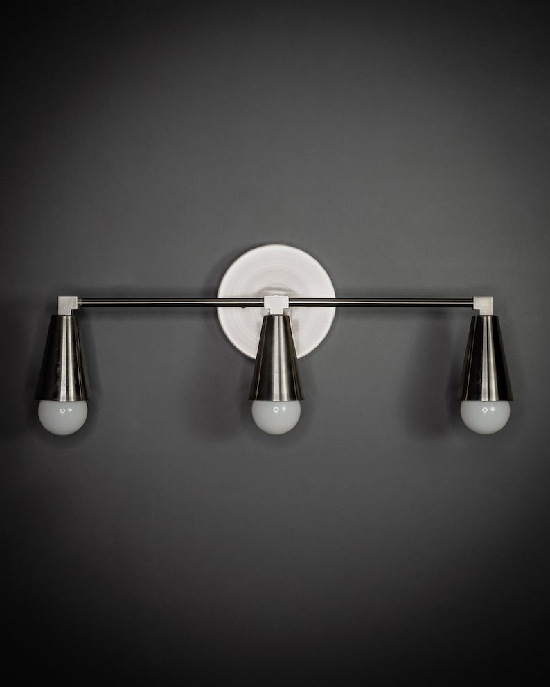 May include: A silver three-light vanity light fixture with a white circular backplate and three cone-shaped shades.