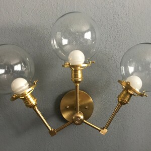 Könnte beinhalten: Eine goldene Wandleuchte mit drei klaren Glaskugeln und freiliegenden Glühbirnen. Die Leuchte hat einen runden Sockel und drei Arme, die sich nach außen erstrecken, um die Kugeln zu halten.