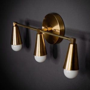 May include: A gold-toned wall sconce with three cone-shaped light fixtures. The sconce has a round base and a straight bar connecting the light fixtures.