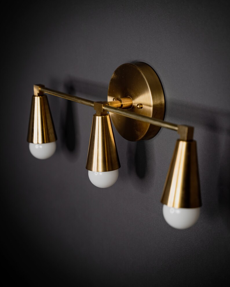 May include: A gold-toned wall sconce with three cone-shaped light fixtures. The sconce has a round base and a straight bar connecting the light fixtures.