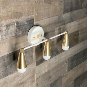 May include: White wall-mounted light fixture with three gold cone-shaped shades and a white metal bar.
