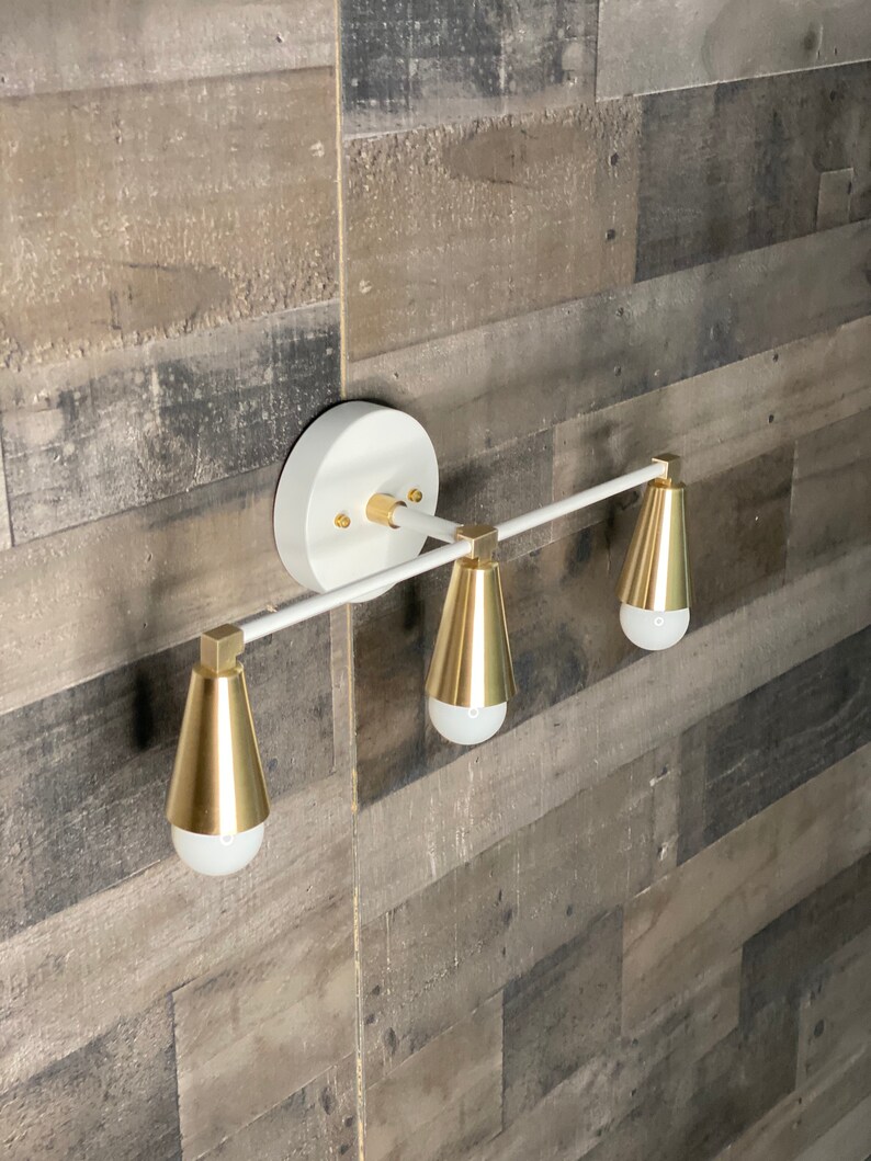 May include: White wall-mounted light fixture with three gold cone-shaped shades and a white metal bar.