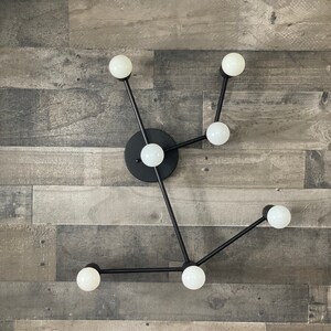May include: A modern black metal light fixture with six white globe light bulbs. The fixture is mounted on a wall with a wood panel background.