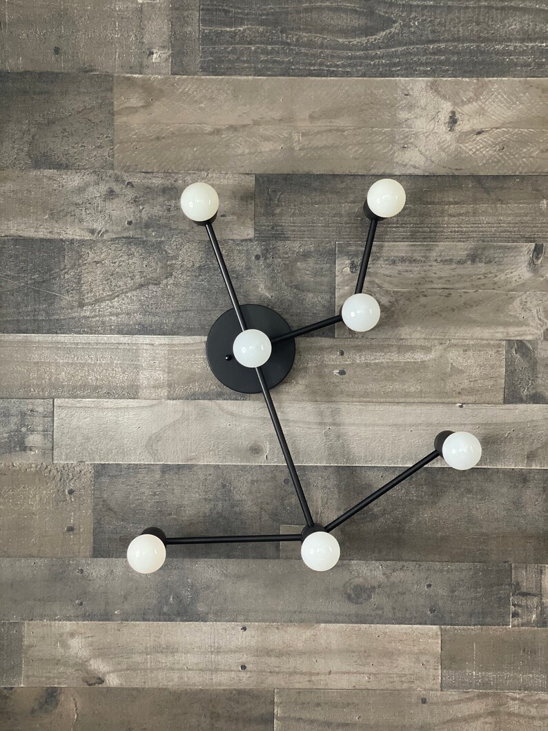 May include: A modern black metal light fixture with six white globe light bulbs. The fixture is mounted on a wall with a wood panel background.