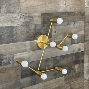May include: Gold metal wall sconce with six light bulbs. The sconce has a geometric design with multiple arms extending from a central point. The light bulbs are all white and are turned on.