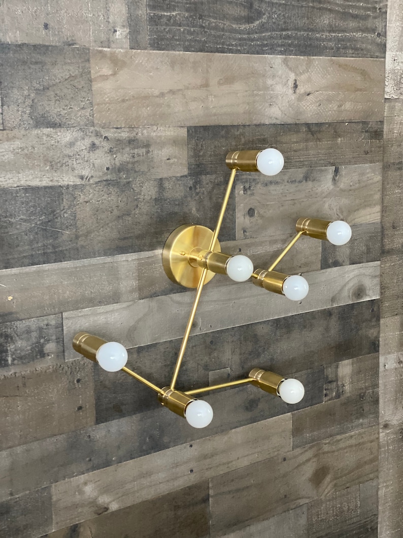 May include: Gold metal wall sconce with six light bulbs. The sconce has a geometric design with multiple arms extending from a central point. The light bulbs are all white and are turned on.