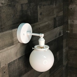 May include: White wall-mounted light fixture with a round white glass shade and a white metal arm.