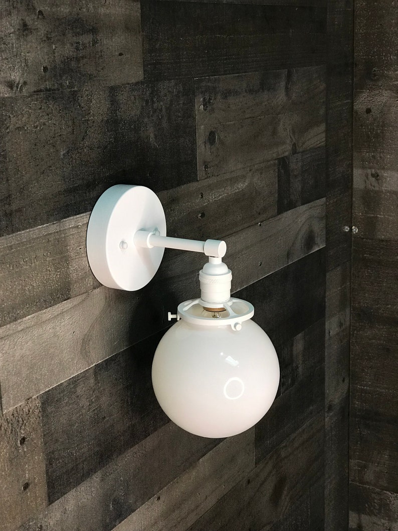 May include: White wall-mounted light fixture with a round white glass shade and a white metal arm.