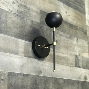 May include: A black and gold wall sconce with a round, black shade and a long, thin arm. The sconce is mounted on a white wall.