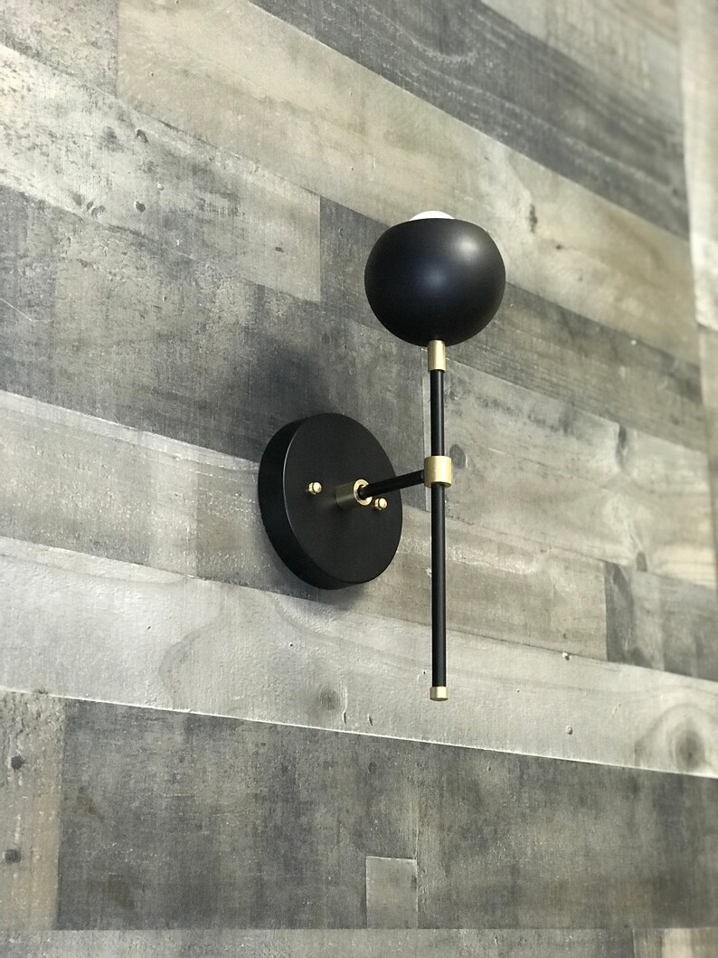 May include: A black and gold wall sconce with a round, black shade and a long, thin arm. The sconce is mounted on a white wall.
