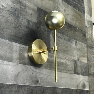 May include: A gold-toned wall sconce with a round, orb-shaped light fixture. The sconce has a simple, modern design and is attached to a wall with a circular base.