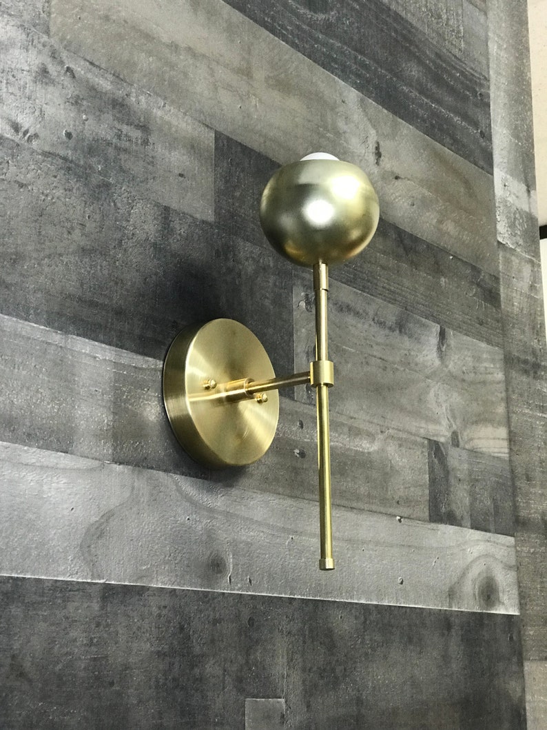 May include: A gold-toned wall sconce with a round, orb-shaped light fixture. The sconce has a simple, modern design and is attached to a wall with a circular base.