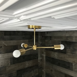 Peut inclure: Un luminaire de plafond en or avec trois bras et trois ampoules blanches. Le luminaire est monté sur un plafond blanc.