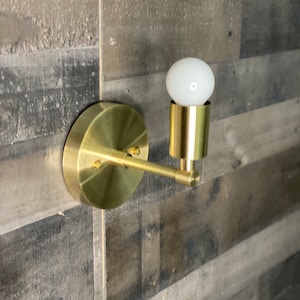 May include: A gold wall sconce with a single light bulb. The sconce has a round base and a single arm that extends out to the side. The light bulb is a standard incandescent bulb.