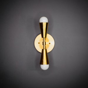 以下が含まれることがあります： 2つの電球が付いた金色の壁掛け照明。照明は丸いベースと2つの円錐形のシェードを持っています。