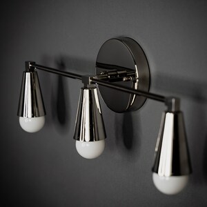 May include: A three-light wall sconce with a polished nickel finish. The sconce has a modern design with three cone-shaped shades.