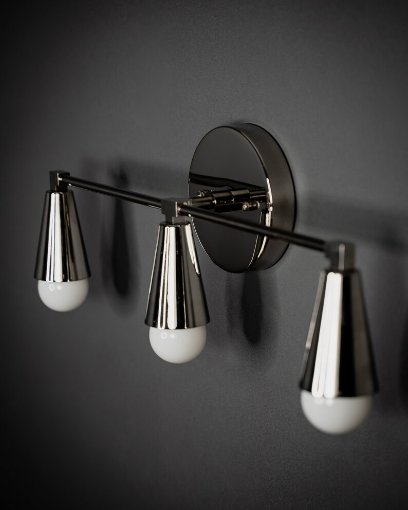 May include: A three-light wall sconce with a polished nickel finish. The sconce has a modern design with three cone-shaped shades.