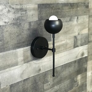 May include: Black modern wall sconce with a round, black shade and a long, thin arm. The sconce is mounted on a white wall.