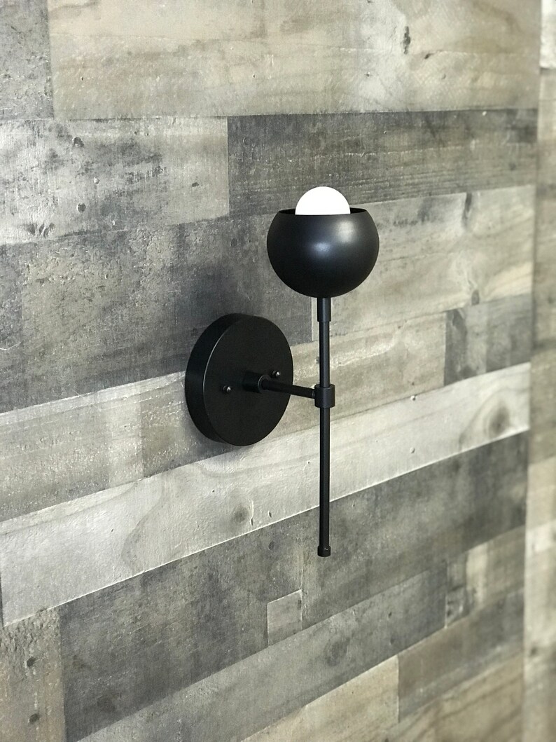 May include: Black modern wall sconce with a round, black shade and a long, thin arm. The sconce is mounted on a white wall.