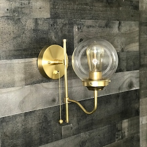 May include: A gold wall sconce with a clear glass globe shade and a pull chain. The sconce is mounted on a wood wall.