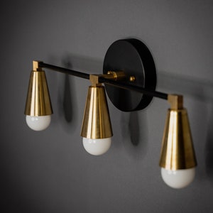 May include: A black and gold wall sconce with three light bulbs. The sconce has a round black base and three gold cone-shaped shades.