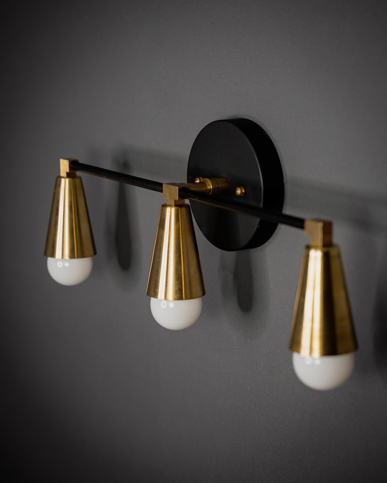 May include: A black and gold wall sconce with three light bulbs. The sconce has a round black base and three gold cone-shaped shades.
