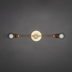 Op de afbeelding: Een gouden wandlamp met drie lampen. De lamp heeft een eenvoudig, modern ontwerp met een rechte stang en ronde lampen.