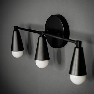 May include: A black wall-mounted light fixture with three cone-shaped shades and exposed bulbs.