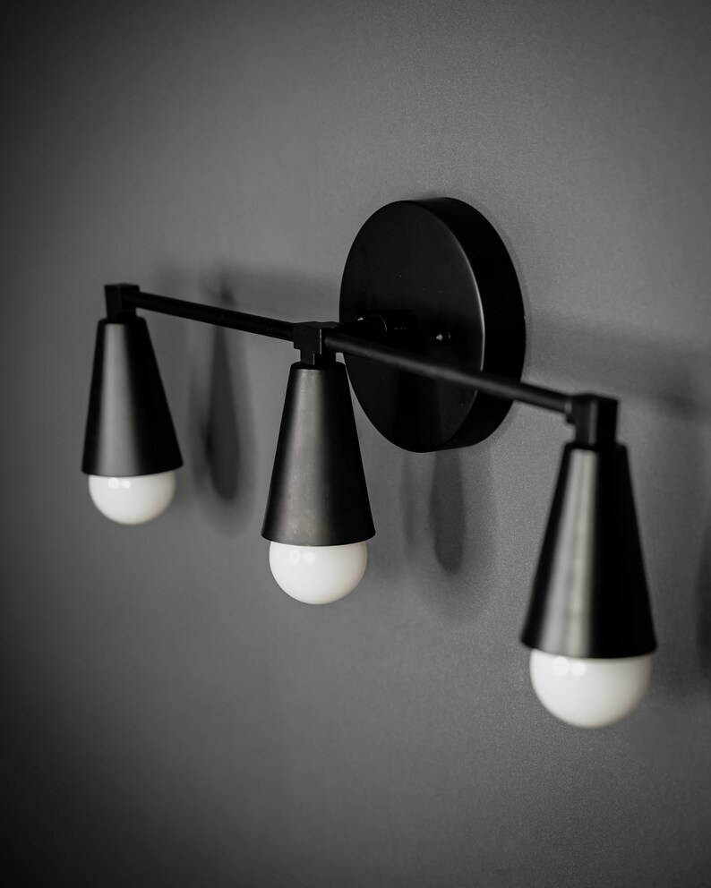 May include: A black wall-mounted light fixture with three cone-shaped shades and exposed bulbs.