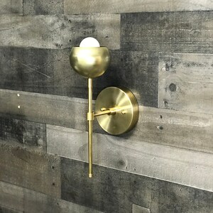 May include: A gold metal wall sconce with a round, globe-shaped light fixture. The sconce has a simple, modern design and is perfect for adding a touch of elegance to any room.