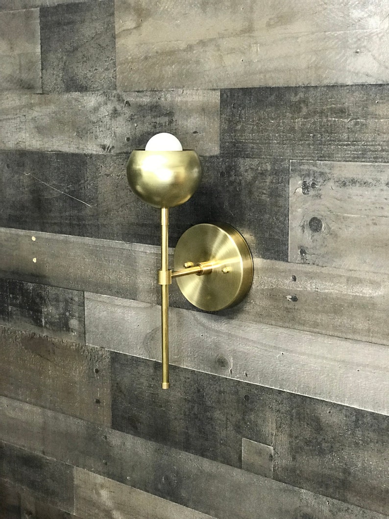 May include: A gold metal wall sconce with a round, globe-shaped light fixture. The sconce has a simple, modern design and is perfect for adding a touch of elegance to any room.