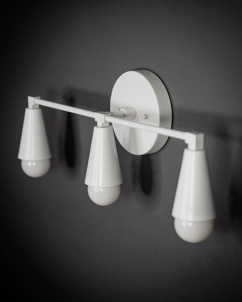 May include: White wall-mounted light fixture with three cone-shaped shades and exposed bulbs.