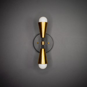 以下が含まれることがあります： 2つの電球が付いた金色と黒色の壁掛け照明。照明は円形のベースと2つの円錐形のシェードを持っています。