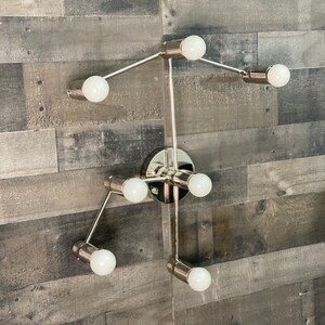 May include: A modern, silver-toned wall sconce with six light bulbs. The sconce has a geometric design with multiple arms extending from a central hub.