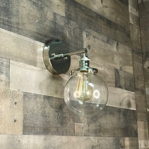 May include: A chrome wall sconce with a clear glass globe shade and a vintage-style light bulb. The sconce is mounted on a wood wall.