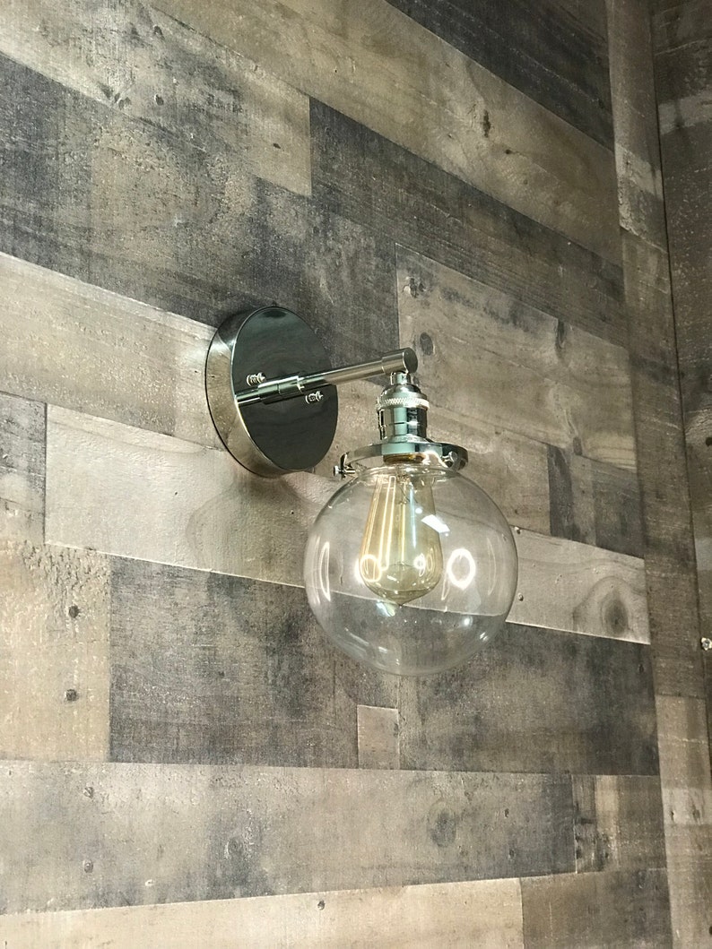 May include: A chrome wall sconce with a clear glass globe shade and a vintage-style light bulb. The sconce is mounted on a wood wall.