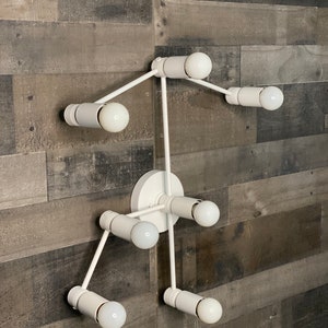 May include: White metal wall sconce with six light bulbs. The sconce has a modern, geometric design with a central hub and arms extending out in different directions.