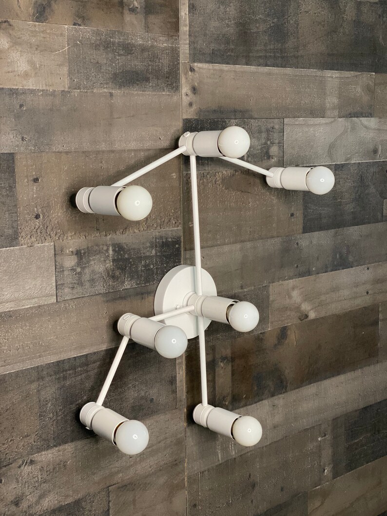 May include: White metal wall sconce with six light bulbs. The sconce has a modern, geometric design with a central hub and arms extending out in different directions.