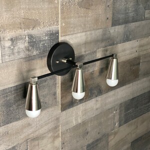 May include: A black and silver three-light vanity light fixture with a modern design. The fixture has a black metal bar with three silver light bulbs.