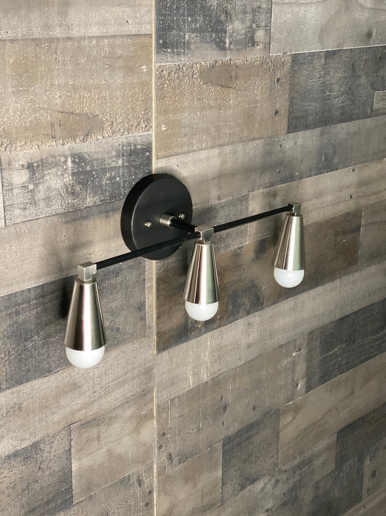 May include: A black and silver three-light vanity light fixture with a modern design. The fixture has a black metal bar with three silver light bulbs.
