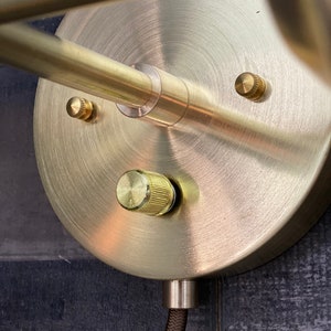 May include: A gold-toned wall sconce with a round base and a single light fixture. The sconce has a brushed metal finish and a cord with a black braided covering.