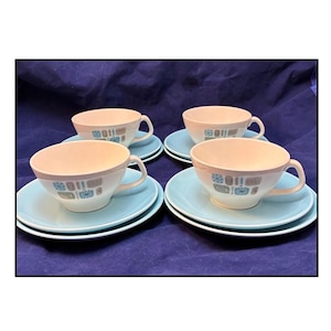 Op de afbeelding: Vier vintage kopjes en schotels. De kopjes zijn gebroken wit met een decoratieve band van blauwe, grijze en bruine geometrische vormen. De schotels zijn lichtblauw van kleur. De set is geplaatst op een donkerblauwe ondergrond.