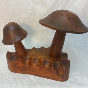 May include: A wooden mushroom-shaped salt and pepper shaker set. The set features two mushroom-shaped shakers, one large and one small, attached to a carved wooden base. The shakers are a warm brown color and have a rustic, handcrafted appearance.