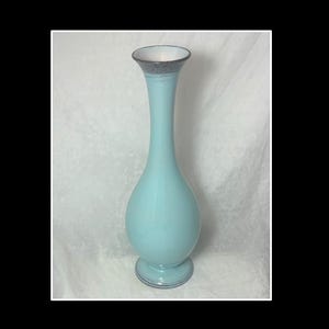 Puede incluir: Un jarrón de cristal alto de color azul claro con una base bulbosa y un cuello fino y alargado. El borde y la base circular presentan un acabado texturizado gris oscuro moteado que contrasta con la superficie lisa y brillante del cuerpo.