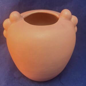 Puede incluir: Un jarrón de cerámica color melocotón con un cuerpo redondeado y una abertura ancha. El jarrón tiene dos protuberancias decorativas y redondeadas a cada lado de la abertura. La forma general recuerda a un corazón.
