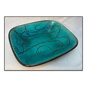 May include: A rectangular ceramic tray with rounded corners, featuring a vibrant turquoise glaze with a dark, uneven finish. The surface is decorated with black, hand-drawn abstract line art consisting of fluid curves and small hatch marks.