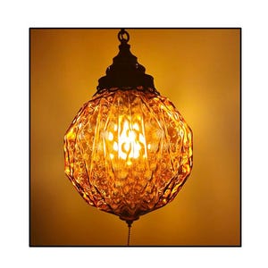 May include: A vintage-style hanging lamp with a textured, amber-colored glass shade. The lamp has a dark metal cap and a pull chain. The light inside the shade is bright, creating a warm glow. The lamp's design is reminiscent of mid-century modern decor.