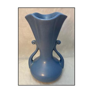 May include: A blue ceramic vase with a flared top and two curved handles. The vase has a rounded base and a smooth, matte finish. The overall shape is reminiscent of a tulip or a trumpet.